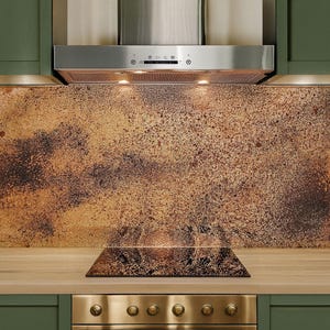 May include: A kitchen scene featuring a stainless steel range hood above a backsplash with a mottled brown and gold pattern. The cabinets are green with gold hardware, and the cooktop is black.