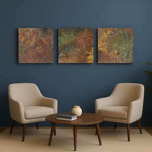 May include: Three square abstract art pieces with a textured, mottled appearance in shades of brown, gold, and green hang above two beige armchairs and a round wooden coffee table. A small vase and a book are on the table.