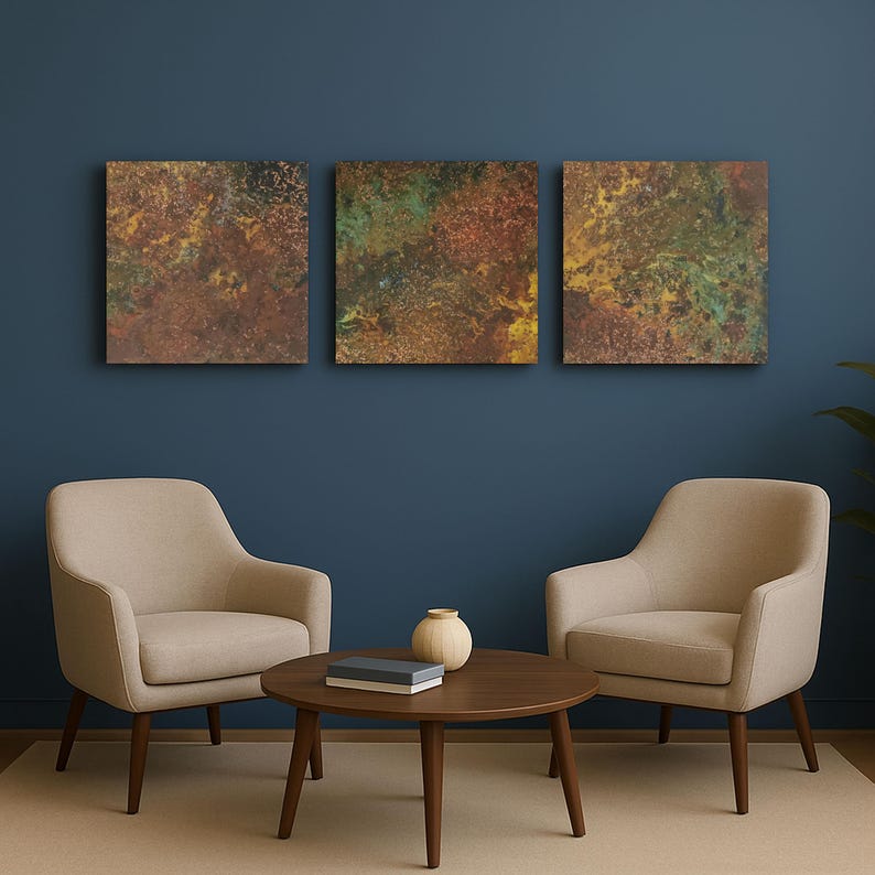 May include: Three square abstract art pieces with a textured, mottled appearance in shades of brown, gold, and green hang above two beige armchairs and a round wooden coffee table. A small vase and a book are on the table.