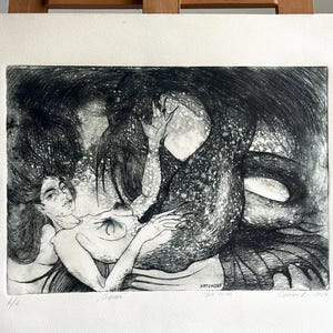 Puede incluir: Un grabado en blanco y negro sobre papel que muestra una figura con cabello largo y fluido abrazando a una gran criatura marina con escamas. La obra incluye el texto manuscrito "1/1", "Ophidia", "etching on paper" y una firma con la fecha "2023".