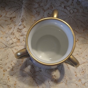 May include: A white porcelain vase with three gold handles. The vase has a gold rim and a blue and white floral design.