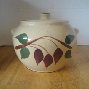 May include: A cream-colored ceramic casserole dish with a matching lid and two side handles. The exterior features a hand-painted design of a brown branch with green leaves and dark red teardrop-shaped berries encircling the center of the pot.