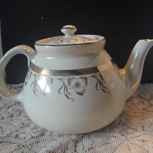May include: Cream-colored teapot with a lid and curved handle. The teapot features a gold band and floral design. The spout is curved and the lid has a small knob. The teapot is sitting on a lace tablecloth.