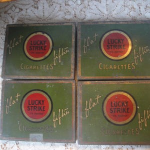 May include: Four vintage green and gold Lucky Strike cigarette tins. Each tin has a red circle with the words "Lucky Strike" and "It's toasted" in white. The tins are labeled "flat fifties" and "cigarettes".