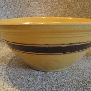 May include: A yellow ceramic mixing bowl with two dark brown bands around the top. The bowl has a slight glaze and is slightly chipped.