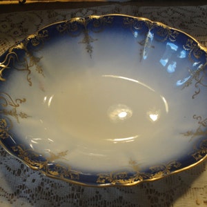 May include: A large oval serving bowl with a white interior and a blue and gold rim. The rim is decorated with a gold floral pattern.