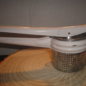 May include: A white metal potato masher with a handle and a perforated metal base. The masher is sitting on a wooden surface.