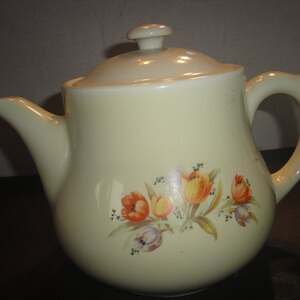 May include: A vintage cream-colored teapot with a floral design featuring orange and yellow tulips. The teapot has a lid with a small knob on top.