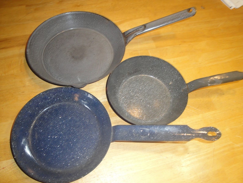 Puede incluir: Tres sartenes vintage de hierro fundido esmaltado. Una es azul con motas blancas, y dos son grises con motas blancas. Las sartenes tienen mangos largos.