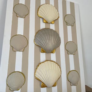 May include: A rectangular wall art piece featuring a white and beige vertical striped background. Nine scallop shells are arranged in a grid pattern, each painted in shades of cream or grey with metallic gold edges on the shell rims.