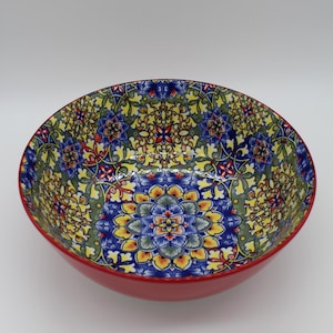 Könnte beinhalten: Eine runde Keramikschüssel mit einem lebendigen Blumenmuster in Blau, Gelb, Grün und Rot. Die Schüssel hat eine einfarbige rote Außenseite und ein detailliertes, symmetrisches Design im Inneren, geeignet zum Servieren von Speisen.
