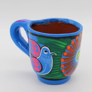 Könnte beinhalten: Eine kleine blaue Keramiktasse mit einem bemalten Design aus einem Vogel, Blumen und Blättern. Die Tasse hat einen braunen Toninnenraum und einen Henkel.