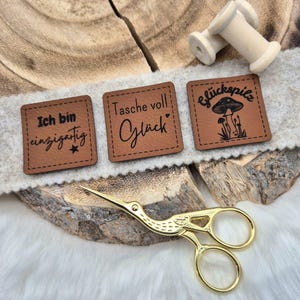 Könnte beinhalten: Drei braune Lederpatches mit schwarzem Text und Designs. Die Patches sind quadratisch und haben genähte Kanten. Der Text auf den Patches lautet "Ich bin einzigartig", "Tasche voll Glück" und "Glückspilz". Der dritte Patch hat ein Pilzdesign.