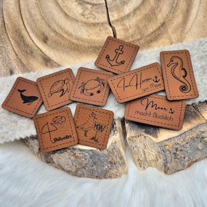 Könnte beinhalten: Neun braune Lederpatches mit verschiedenen maritimen Designs und Texten. Die Designs beinhalten ein Seepferdchen, einen Krebs, eine Schildkröte, einen Anker, einen Wal, eine Möwe, einen Regenschirm und den Text "Im Herzen am Meer" und "Meer macht Glücklich".