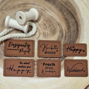 Könnte beinhalten: Sechs braune Leder-Etiketten mit schwarzem Text. Die Etiketten haben verschiedene Sprüche, darunter "Einzigartig ist viel besser als perfekt", "Konfetti im Herzen", "Be Happy", "Do what makes you Happy", "Frech Wild Wunderbar" und "Wirbelwind".