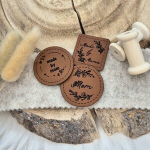 Könnte beinhalten: Drei braune Leder-Etiketten mit genähten Kanten. Die Etiketten haben Blumendesigns und den Text "Made by Mama", "Made by Mom" und "Mom".