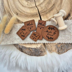 Könnte beinhalten: Fünf braune Leder-Etiketten mit dem Text "Made by Oma" in verschiedenen Schriftarten und Designs. Die Etiketten befinden sich auf einer Holzoberfläche mit einer weißen, flauschigen Oberfläche im Hintergrund.