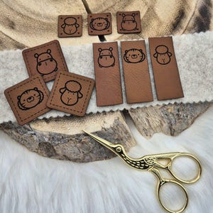 Könnte beinhalten: Ein Set aus acht braunen Leder-Patches mit verschiedenen Tiermotiven, darunter ein Flusspferd, ein Tiger, ein Schaf und eine Katze. Die Patches sind an den Rändern vernäht und haben einen rustikalen, Vintage-Look. Eine goldene Schere ist ebenfalls im Bild enthalten.