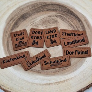 Könnte beinhalten: Neun braune Leder-Patches mit schwarzem Text und Bildern. Die Patches sagen "Stadt Kind", "Dorf Kind", "Land Kind", "Stadtkind", "Landkind", "Dorfkind", "Küstenkind", "Schulkind" und "Schulkind".
