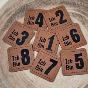 Könnte beinhalten: Acht braune Lederquadrate mit schwarzen Nähten und schwarzen Zahlen von 1 bis 8. Jedes Quadrat trägt den Text "Ich Bin" in Schwarz.