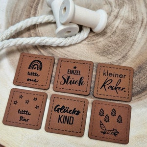 Könnte beinhalten: Sechs braune Leder-Geschenk-Etiketten mit schwarzem Text und Designs. Die Etiketten zeigen Sätze wie "little one", "Glücks Kind" und "kleiner Racker".