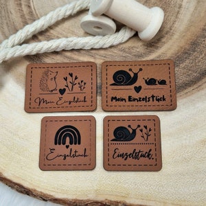 Könnte beinhalten: Vier braune Leder-Etiketten mit schwarzen Nähten und Designs. Die Etiketten zeigen einen Igel, eine Schnecke, einen Regenbogen und eine Schnecke mit einem Herzen. Der Text auf den Etiketten lautet "Mein Einzelstück" und "Einzelstück".