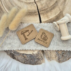 Könnte beinhalten: Zwei braune Lederpatches mit genähten Kanten. Ein Patch zeigt einen Kran und den Text "Baustelle". Der andere Patch zeigt einen Bagger und den Text "BAUSTELLE".
