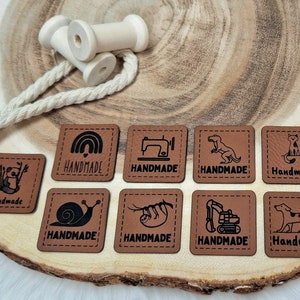 Könnte beinhalten: Neun braune Leder-Etiketten mit schwarzen Nähten und verschiedenen Illustrationen sowie dem Wort "Handmade". Die Illustrationen umfassen einen Regenbogen, eine Nähmaschine, einen Dinosaurier, eine Katze, eine Schnecke, ein Faultier, einen Bagger und einen Hund.