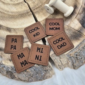 Könnte beinhalten: Sechs braune Leder-Patches mit genähten Kanten und schwarzem Text. Die Patches lauten "Cool Kid", "Cool Mom", "Cool Dad", "Pa Pa", "Ma Ma" und "Mi Ni".