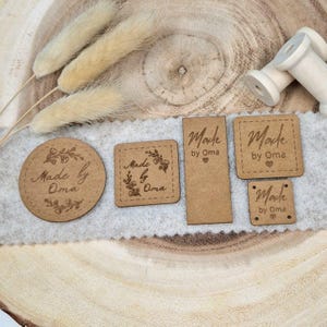 Könnte beinhalten: Vier braune Holzetiketten mit dem Text "Made by Oma" und einem kleinen Blumenmuster. Die Etiketten haben verschiedene Formen und Größen, darunter ein Kreis, ein Quadrat und ein Rechteck.