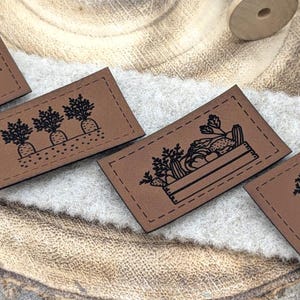 Könnte beinhalten: Vier rechteckige braune Lederetiketten mit schwarzen Illustrationen zu Gartenthemen. Die Etiketten haben genähte Kanten und zeigen Blumen, Karotten, eine Gemüsekiste und eine Schubkarre mit Kürbissen.