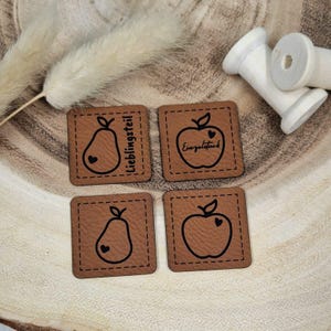 Könnte beinhalten: Vier braune Leder-Patches mit schwarzen Nähten und Designs. Zwei Patches zeigen eine Birne mit einem Herzen, einer sagt "Lieblingsteil" und der andere hat keinen Text. Die anderen beiden Patches zeigen einen Apfel mit einem Herzen, einer sagt "Eingepackt" und der andere hat keinen Text.