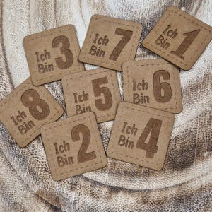 Könnte beinhalten: Neun braune quadratische Karten mit abgerundeten Ecken und weißen Nähten an den Rändern. Jede Karte hat eine Zahl von 1 bis 8 in Braun auf der Vorderseite gedruckt. Der Text "Ich Bin" ist über jeder Zahl gedruckt.