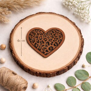 Könnte beinhalten: Eine herzförmige Holzdekoration mit Leopardenmuster, 4 cm hoch, wird auf einer Holzscheibe präsentiert. Das Herz ist braun mit einem schwarzen Leopardenmuster. Die Holzscheibe hat eine natürliche Holzmaserung.