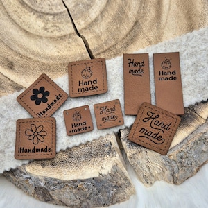 Könnte beinhalten: Sortiment aus braunen Leder-Etiketten mit dem Text "Hand made" in verschiedenen Schriftarten und Stilen. Einige Etiketten zeigen ein Marienkäfer-Design, während andere ein Blumendesign haben.
