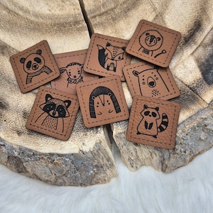 Könnte beinhalten: Neun braune Lederquadrate mit genähten Kanten, jedes mit einer anderen Tierillustration in Schwarz: ein Panda, ein Schaf, ein Fuchs, ein Bär, ein Igel, ein Waschbär und zwei weitere Waschbären.