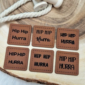 Könnte beinhalten: Sechs braune Leder-Patches mit dem Text "Hip Hip Hurra" in schwarzer Schrift. Die Patches sind an den Rändern vernäht.