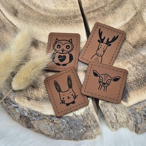 Könnte beinhalten: Vier braune Leder-Patches mit schwarzen, gesteppten Kanten. Jeder Patch zeigt ein anderes Tier: eine Eule, ein Reh, ein Kaninchen und ein Kitz.