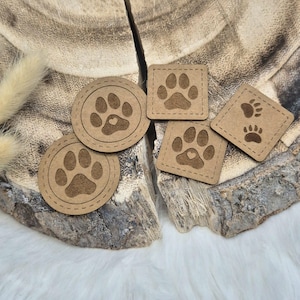 Könnte beinhalten: Vier braune Leder-Pfotenabdruck-Patches mit einem Herz in der Mitte jedes Pfotenabdrucks. Die Patches befinden sich auf einer Holzoberfläche.