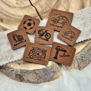 Könnte beinhalten: Sechs braune Lederpatches mit schwarzen, gesteppten Kanten und verschiedenen Designs. Die Patches zeigen ein Fahrrad, ein Auto, ein Feuerwehrauto, einen Baukran, einen Fußball und ein Baufahrzeug. Die Patches haben deutschen Text darauf, darunter "Tutu", "Selbstgemacht", "Bike on Tour", "Tatu Tata" und "Baustelle".