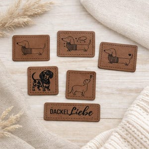 Könnte beinhalten: Eine Sammlung brauner Lederpatches mit schwarzen Dackel-Illustrationen. Die Patches sind rechteckig und quadratisch, mit genähten Kanten. Ein Patch trägt den Text "DACKEL Liebe". Die Hunde werden in verschiedenen Posen dargestellt.