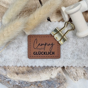 Könnte beinhalten: Ein braunes Leder-Patch mit dem Text "Camping macht glücklich" in schwarzer Schrift. Das Patch ist mit einem goldenen Büroklammer an einer weißen Oberfläche befestigt.