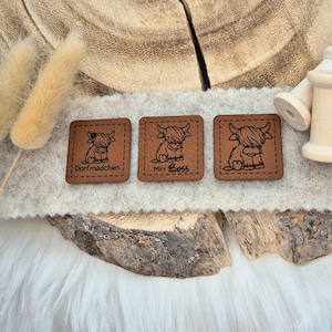 Könnte beinhalten: Drei braune Lederpatches mit schwarzem Umriss einer Highland-Kuh. Die Patches haben den Text "Dorfmadchen", "Mini Boss" und ein drittes Patch nur mit der Kuh.