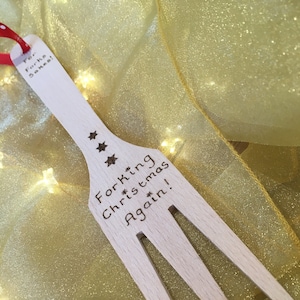 May include: Wooden fork-shaped Christmas ornament with the text "Forking Christmas Again!" and "For Forks Sakes!" etched into the wood. The ornament has three star designs and a red ribbon for hanging. The background is a gold fabric.
