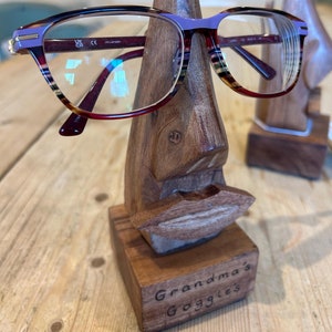 Puede incluir: Un soporte de madera con forma de cara con un par de gafas moradas y marrones apoyadas en la nariz. El soporte tiene una base de madera con el texto "Grandma's Goggles" grabado en ella.