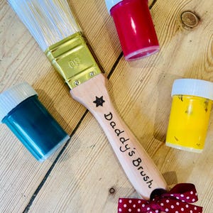 May include: A paintbrush with white bristles, a gold-toned metal ferrule marked 30, and a light wood handle engraved with a star and the text Daddy's Brush. A maroon ribbon with white polka dots is tied to the handle, alongside three small paint pots.