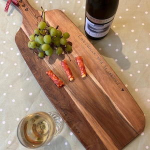 Könnte beinhalten: Ein Holz-Servierbrett mit Griff und dem eingravierten Text "Eat, drink and be retired..." und "We'll miss you from your finance crew xxx". Auf dem Brett liegen eine Flasche Weißwein, Trauben und Salami.