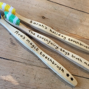 Könnte beinhalten: Drei Holz-Zahnbürsten mit bunten Borsten. Die Zahnbürsten haben personalisierte Botschaften: "Sarah's holiday toothbrush", "The toothfairy says brush more!" und "Ava's magic toothbrush".
