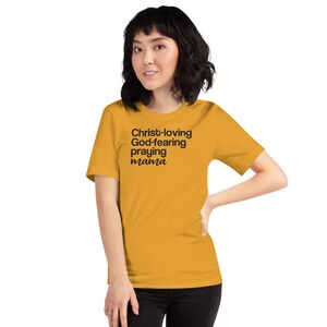 May include: Mustard yellow t-shirt featuring the black text "Christ-loving God-fearing praying mama". The shirt is a short-sleeved crew neck style.
