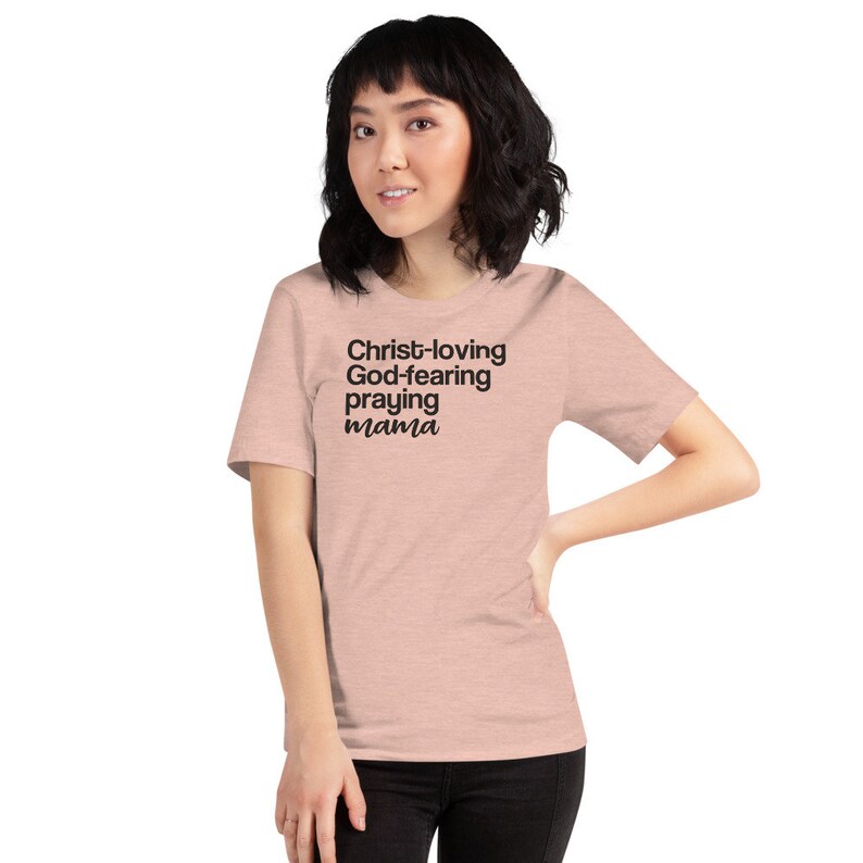 May include: A pale pink t-shirt with the text "Christ-loving, God-fearing, praying mama" in black. The short-sleeved shirt is made of a soft fabric. The model is wearing the shirt.